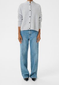 Hellgraue, gerippte Strickjacke mit hohem Kragen und dunklen Knöpfen, kombiniert mit blauen, weit geschnittenen Jeans und schwarzen spitzen High Heels.