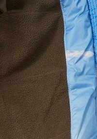 Brauner Fleece-Stoff neben blauer Nylonware, mit glatter Textur und weichem Griff, einer genähten Kante und einem subtilen Wolkenmuster im blauen Abschnitt.