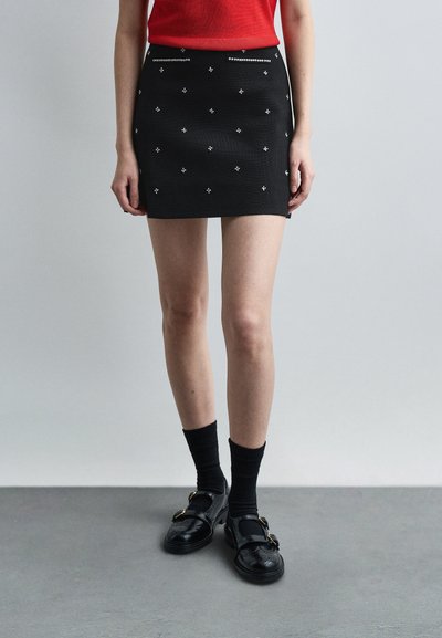 Femme portant une mini jupe noire avec des ornements argentés, des chaussettes noires, des chaussures noires à boucle, et un haut rouge sans manches, debout sur un sol gris.