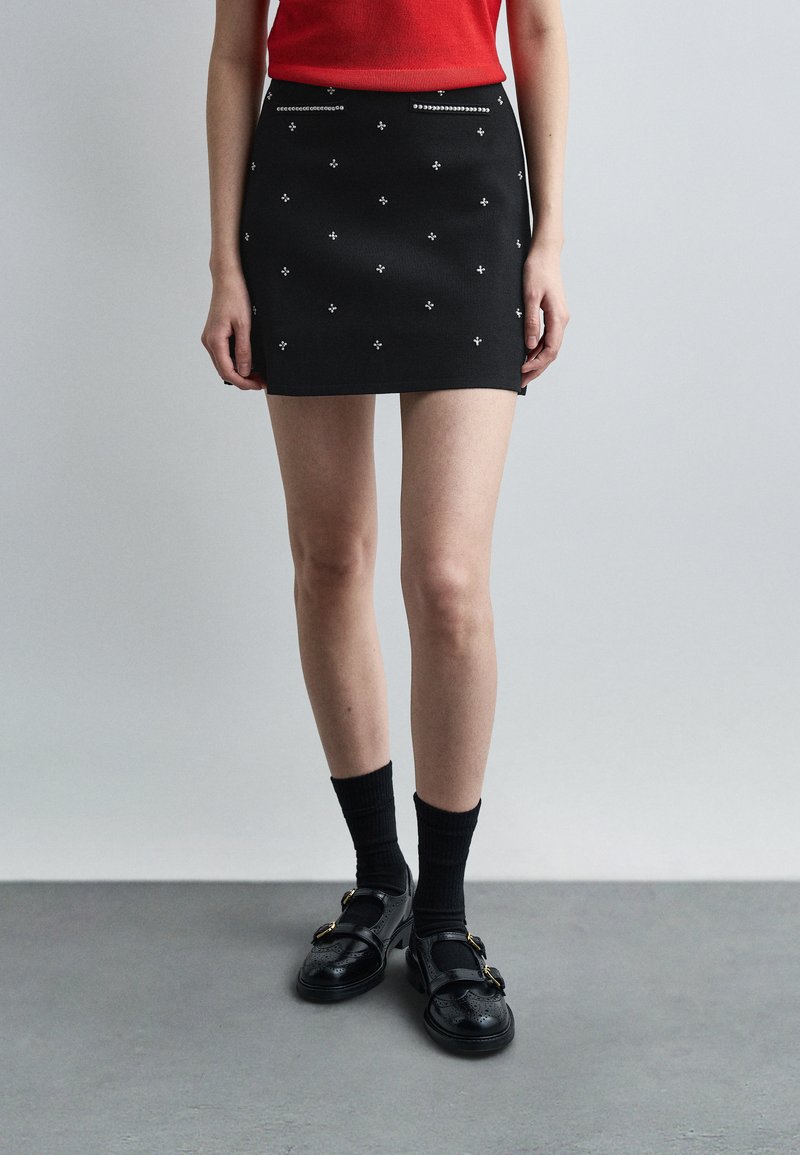 Γυναίκα που φοράει μαύρη μίνι φούστα με ασημένιες διακοσμήσεις, μαύρες κάλτσες, μαύρα παπούτσια με αγκράφες και κόκκινο αμάνικο τοπ, στέκεται σε γκρι πάτωμα.