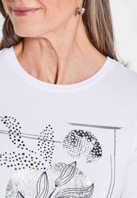 Weißes T-Shirt mit schwarzen und silbernen abstrakten Blumen- und Punktmustern. Rundhalsausschnitt, weicher Stoff, enganliegende Passform.