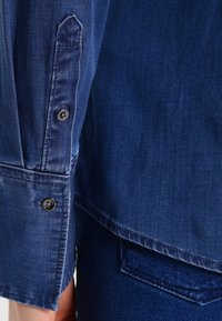 Denimskjorta i mörkblått, med en knappad manschett med två svarta knappar och en mjuk tygtextur. Synliga sömmar vid ärmen.