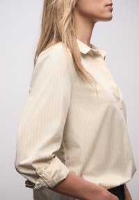 Jasnobeżowa koszula z kołnierzykiem z subtelnymi białymi pionowymi paskami, podwijanymi rękawami i zapięciem na guziki, o gładkiej fakturze.
