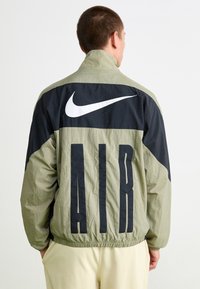 Nike jakna s svetlobno težko, gubano tkanino, z zgornjim delom črne barve, velikimi črnimi črkami "AIR" in izrazitim belim logotipom.