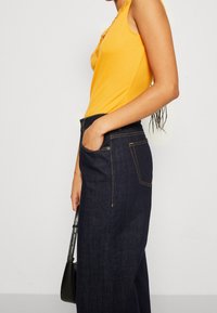 Gelbes geripptes Tanktop mit dekorativen Spitzendetails, kombiniert mit hochtaillierten dunkelblauen Jeanshosen mit orangefarbener Naht und Seitentaschen.