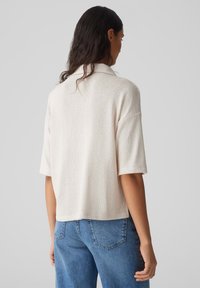 Cremfarbener Pullover mit kurzen Ärmeln, lockerer Passform und strukturiertem Strickmuster, kombiniert mit blauen hochgeschnittenen Jeans. Rückansicht angezeigt.