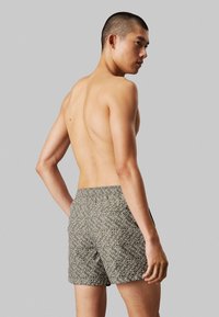 Costumi da bagno da uomo in un tessuto verde oliva scuro con un motivo testuale bianco. Design con elastico in vita e lunghezza sopra il ginocchio.