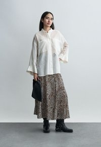 Γυναίκα που φοράει διάφανη λευκή πουκάμισο με κουμπιά, φούστα με λεοπάρ print, μαύρες μπότες με αλυσίδα, κρατώντας ένα μαύρο τσαντάκι μπροστά σε απλό φόντο.