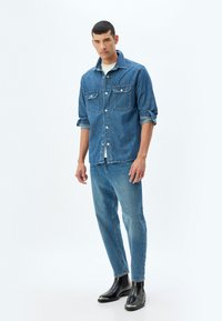 Camisa de denim en azul oscuro con dos bolsillos en el pecho, cierre de botones en la parte delantera, mangas remangadas; combinada con pantalones vaqueros de azul claro y botas negras hasta el tobillo.