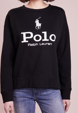 Person iført en sort sweatshirt med hvid Polo Ralph Lauren-logo og blå jeans foran en ensfarvet lys baggrund.