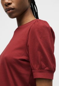 Γυναίκα που φοράει ένα κοντό κόκκινο πουκάμισο με φουσκωτά μανίκια και σφιχτά μανσέτες, με πλεξούδες στα μαλλιά, εμφανιζόμενη από τους ώμους και πάνω, μπροστά σε απλό φόντο.