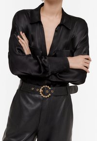 Blusa nera in satin con scollatura a V profonda e maniche lunghe, abbinata a pantaloni in pelle nera a vita alta e una cintura con dettagli dorati.