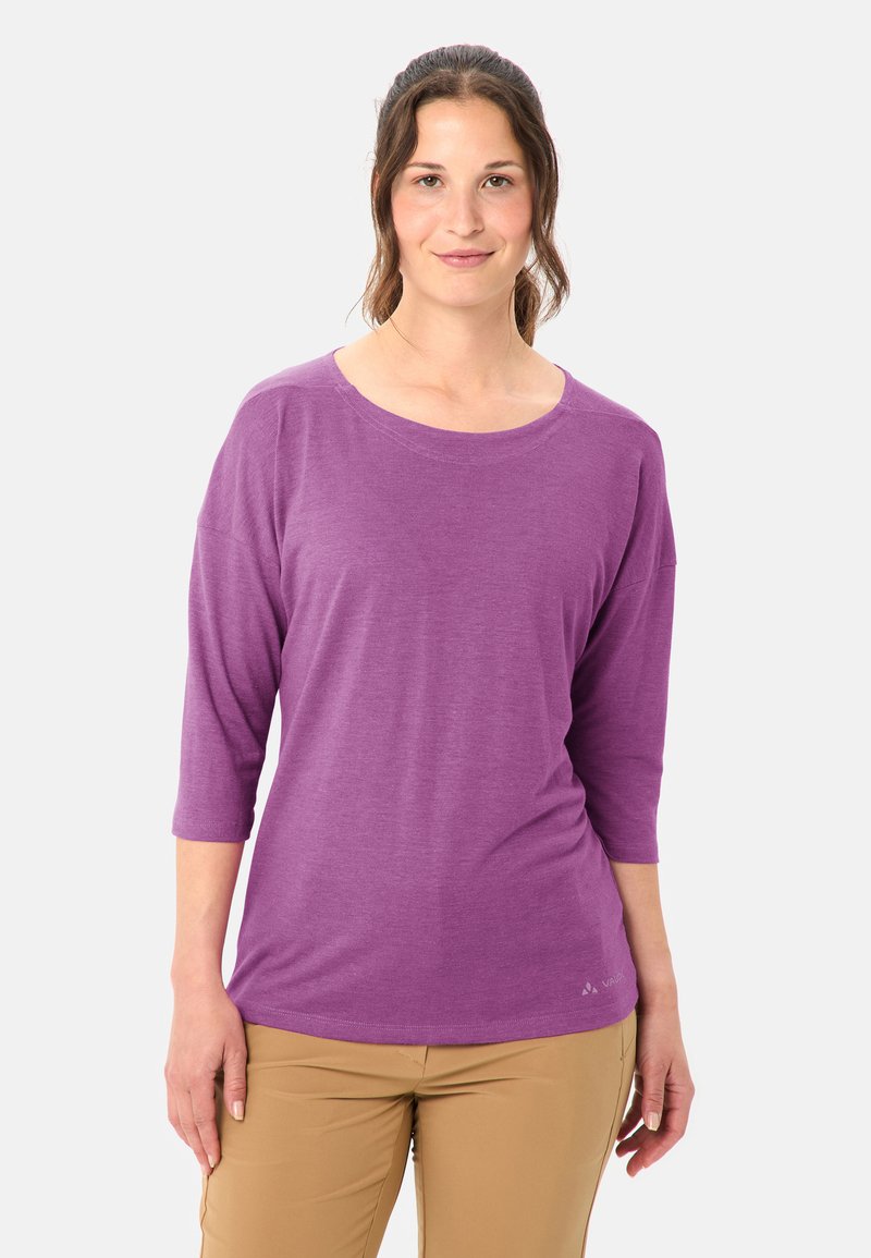Vrouw met bruin haar in een knot draagt een paarse top met driekwart mouwen en beige broek tegen een eenvoudige lichte achtergrond.