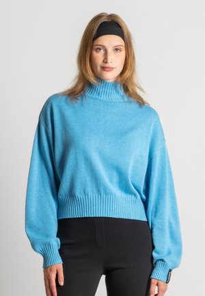 Hellblauer, übergroßer Rollkragenpullover mit geripptem Saum und Bündchen, langen Ärmeln und strukturierter Oberfläche. Kombiniert mit schwarzen Hosen.
