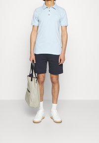 Polo shirt azzurro chiaro con maniche corte, pantaloni shorts blu scuro, sneakers bianche e una borsa tote grigia con manici neri e motivo interno.