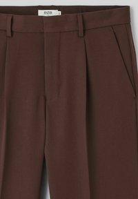 Pantalons pleats marron avec une texture lisse, dotés de poches latérales et de passants de ceinture. L'étiquette indique "B Zeb City" visible près de la taille.