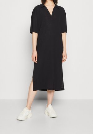 Person iført en midt-lang sort kjole med krave og korte ærmer, kombineret med hvide sneakers, stående mod en ensfarvet lys baggrund.