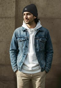 Black knitted beanie with ribbed texture; denim jacket with front pockets; white hoodie underneath; beige pants; standing against a neutral background.