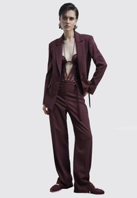 Completo a quadri bordeaux con pantaloni a gamba larga e blazer sartoriale sopra un body in pizzo. Il tessuto ha una finitura testurizzata e un leggero lucido.