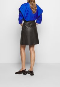 Jupe en cuir noir à coupe ajustée, associée à une chemise bleue aux épaules structurées. Des chaussures plates marron complètent la tenue.