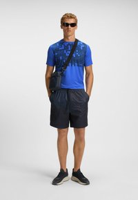 Blå kortærmet skjorte med et sort rutemønster, parret med mørke shorts og sorte sneakers. En lille crossover taske bæres.