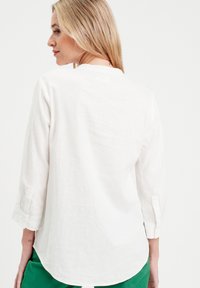 Chemise en lin blanc à manches trois-quarts, dotée d'une coupe décontractée et d'un détail de bouton sur les poignets, présentée sur un fond clair.