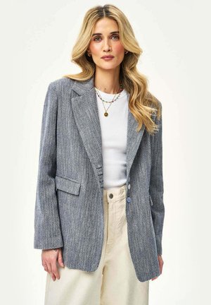 Kobieta z długimi blond włosami ubrana w szarą marynarkę w jodełkę, biały top, kremowe spodnie oraz warstwowe naszyjniki na jednolitym tle.