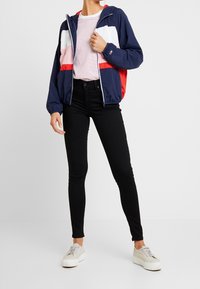 Person som bär en marinblå, röd, rosa och vit sweatshirt med dragkedja, vit skjorta med röda ränder, svarta skinny jeans och vita plattformssneakers.