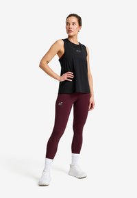 Woman in sleeveless black top and burgundy leggings standing with hand on hip, wearing white sneakers and socks against a plain background.