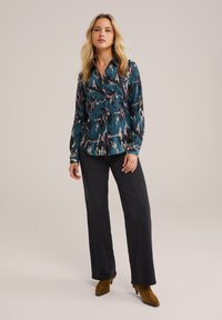 Camicia blu con bottoni, decorata con un motivo astratto multicolore, abbinata a pantaloni larghi e scuri e stivaletti alla caviglia in camoscio marrone.