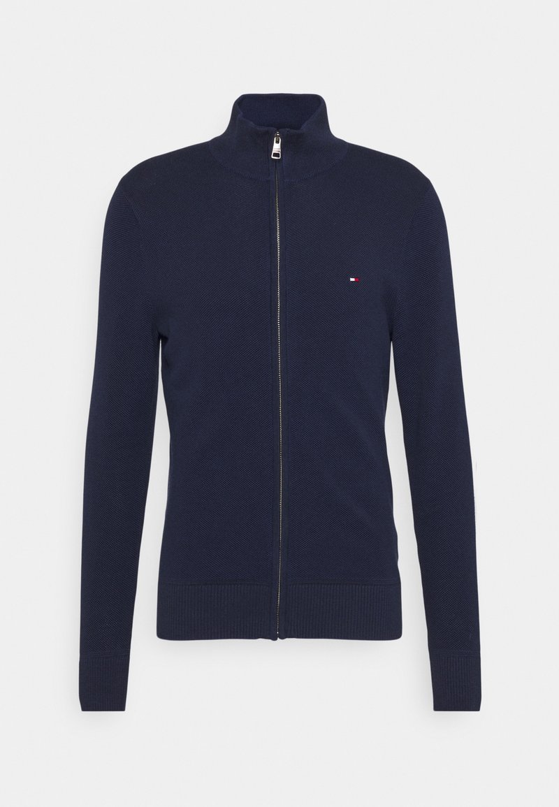 Pull en tricot bleu marine à fermeture éclair intégrale, col montant, manches longues, poignets côtelés, et petit logo rouge, blanc et bleu sur le côté gauche de la poitrine.