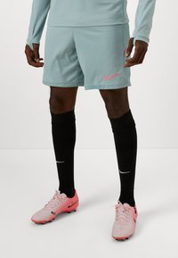 Nike vaaleansiniset urheilushortsit, joissa on vaaleanpunainen logo, yhdistettynä mustiin urheilusukkiin ja vaaleanpunaisiin jalkapallokengiin, joissa on mustia yksityiskohtia.