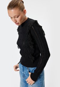 Maglione nero lavorato a maglia con texture a coste, rifinitura in pizzo su spalle e maniche, collo rotondo e polsini aderenti, abbinato a jeans azzurri chiari.