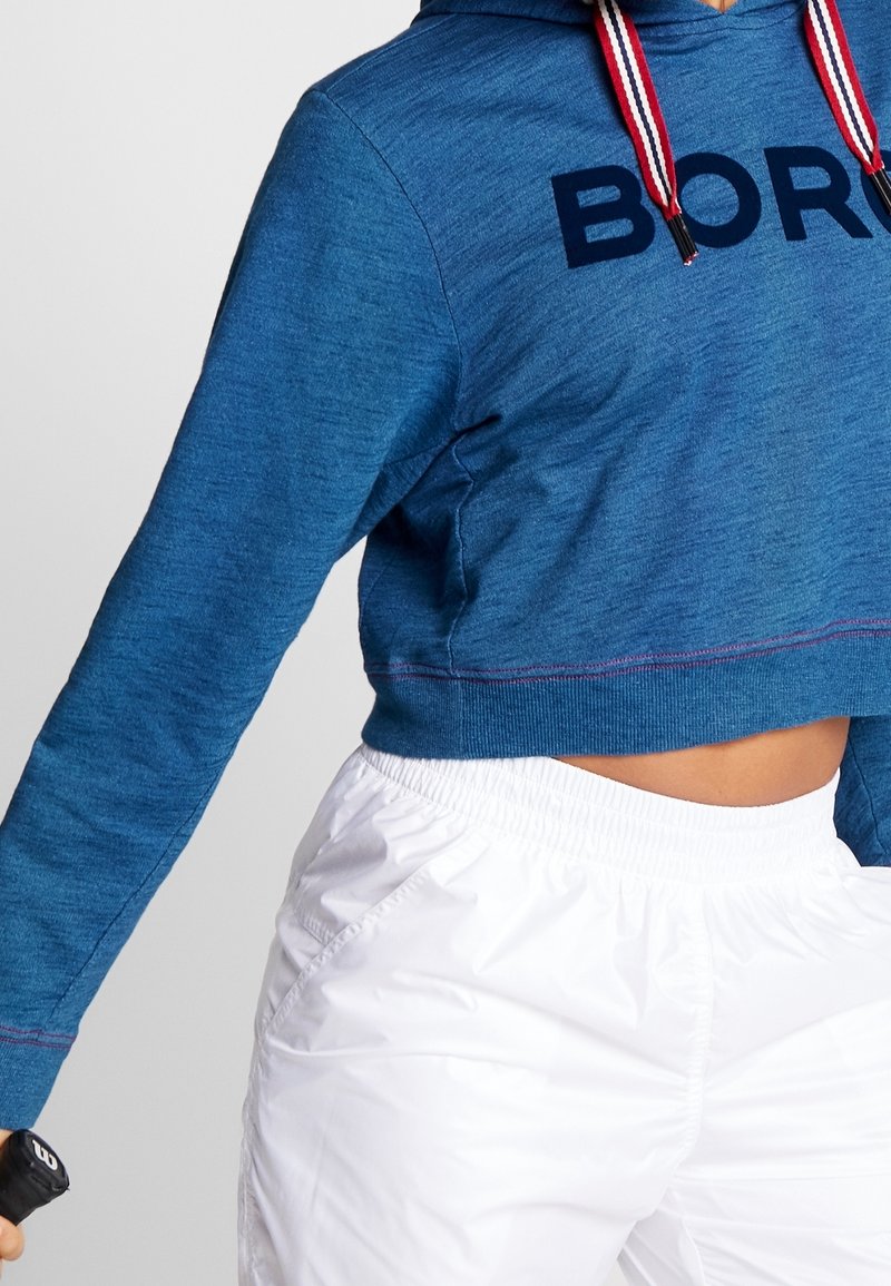 Cropped hoodie blu con scritta navy "BORN," dotato di cappuccio con lacci e cuciture a contrasto. Abbinato a pantaloni corti bianchi con vita elastica.