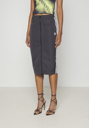 Personne portant une jupe noire Adidas zippée avec des rayures blanches, un crop top vert à motifs, et des sandales noires à talons hauts avec des lanières.