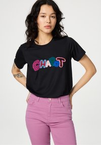 Zwarte t-shirt met kleurrijke geborduurde tekst "CHAPOT." Gepaarde met roze jeans, die een gladde stof en een aangesloten ontwerp tonen.