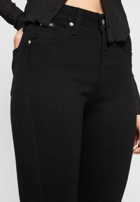 Jeans de denim pretos com uma textura suave, cinco bolsos e acessórios em prata, com um corte justo e detalhes de costura subtis.