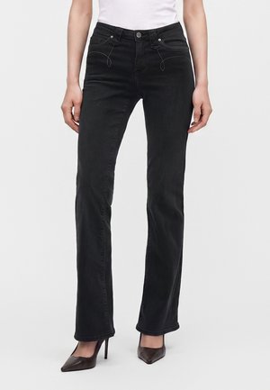 Γυναίκα που φοράει μαύρο τζιν bootcut με ραμμένες λεπτομέρειες μπροστινής τσέπης και σκούρα ψηλοτάκουνα μυτερά παπούτσια σε λευκό φόντο.