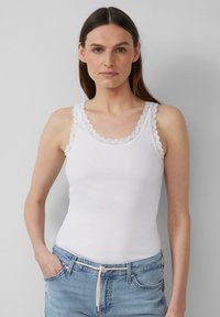 Witte ribbel tanktop met een gegolfde kantafwerking langs de halslijn en de bandjes. Gecombineerd met lichtblauwe jeans, handen in de zakken.