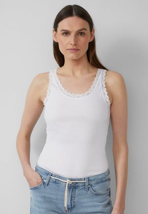 White ribbed tank top with scalloped lace trim along the neckline and straps. Paired with light blue jeans, hands in pockets.