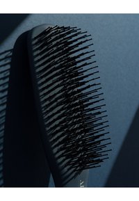 Svart hårbørste med et flatt, avrundet håndtak og flere fine plastbust, som kaster skygger på en mørkeblå bakgrunn.