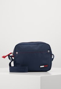 Námořnická nylonová křížová taška s červenými akcenty, zipovým uzávěrem, nastavitelným popruhem a štítkem s logem v modré, bílé a červené barvě. Texturovaný povrch.