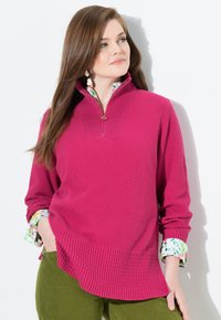 Donna con lunghi capelli castani che indossa un pullover rosa a trama sopra una camicia floreale e pantaloni verdi, in posa con una mano in tasca.