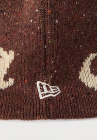 Brązowa dziana czapka z wielokolorowymi kropkami, z kremowym wzorem logo i małą białą wyszywaną metką na daszku.