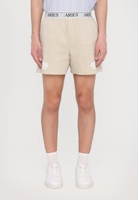 Pantaloni corti beige con una vita elastica regolabile, caratterizzati da un logo "ARIES" in nero. Il lato sinistro presenta ricami floreali bianchi. Indossati con sneakers bianche.