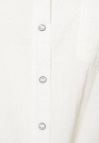 Camisa de cuello blanco con una tela texturizada, que presenta seis botones en la parte delantera con la marca "ONLY" en el botón superior.