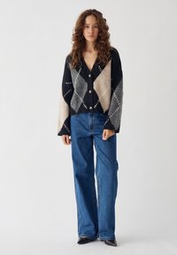 Młoda kobieta z kręconymi włosami, ubrana w czarno-szaro-beżowy sweter w romby i niebieskie dżinsy o szerokich nogawkach, stojąca na tle jednolitego tła.