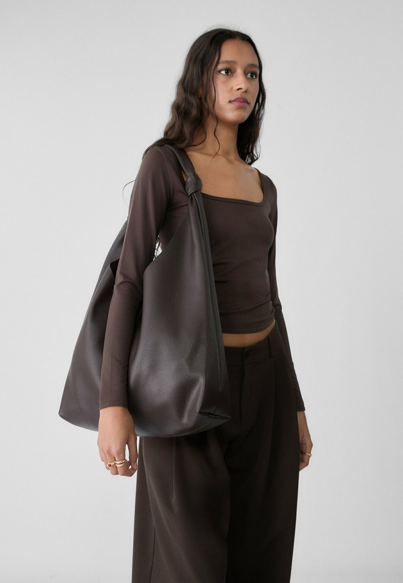 Brown leather hobo bag with a soft texture, rounded shape, and a knotted handle detail, carried over the shoulder against a light background.