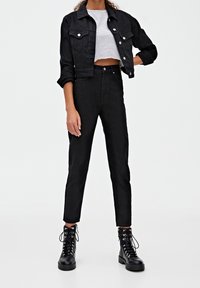 Zwarte denim jack met twee borstzakken, gedragen over een grijs gestreept crop top. Zwarte high-waisted jeans en zwarte veterschoenen maken de look compleet.