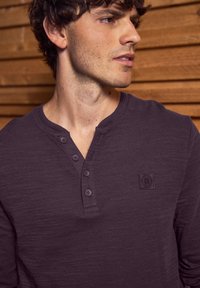 Dark purple long-sleeve henley shirt featuring a textured fabric, four buttons down the front, and a small embroidered logo on the chest.